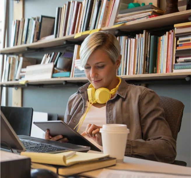 Woman sitting at a desk in front of a computer wearing headphones while working on a tablet